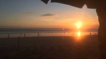 A beach scene with the sun setting over the ocean, silhouettes of people on the sand, and a clear sky.