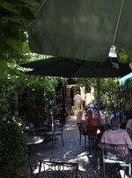 Café al aire libre con mesas y sillas bajo un paraguas grande, rodeado de follaje.