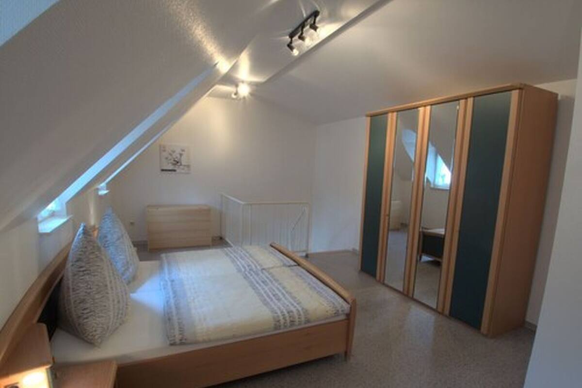 A bedroom with a sloped ceiling, a bed with a wooden frame, a bedside table with a lamp, and a wardrobe with mirrored doors.