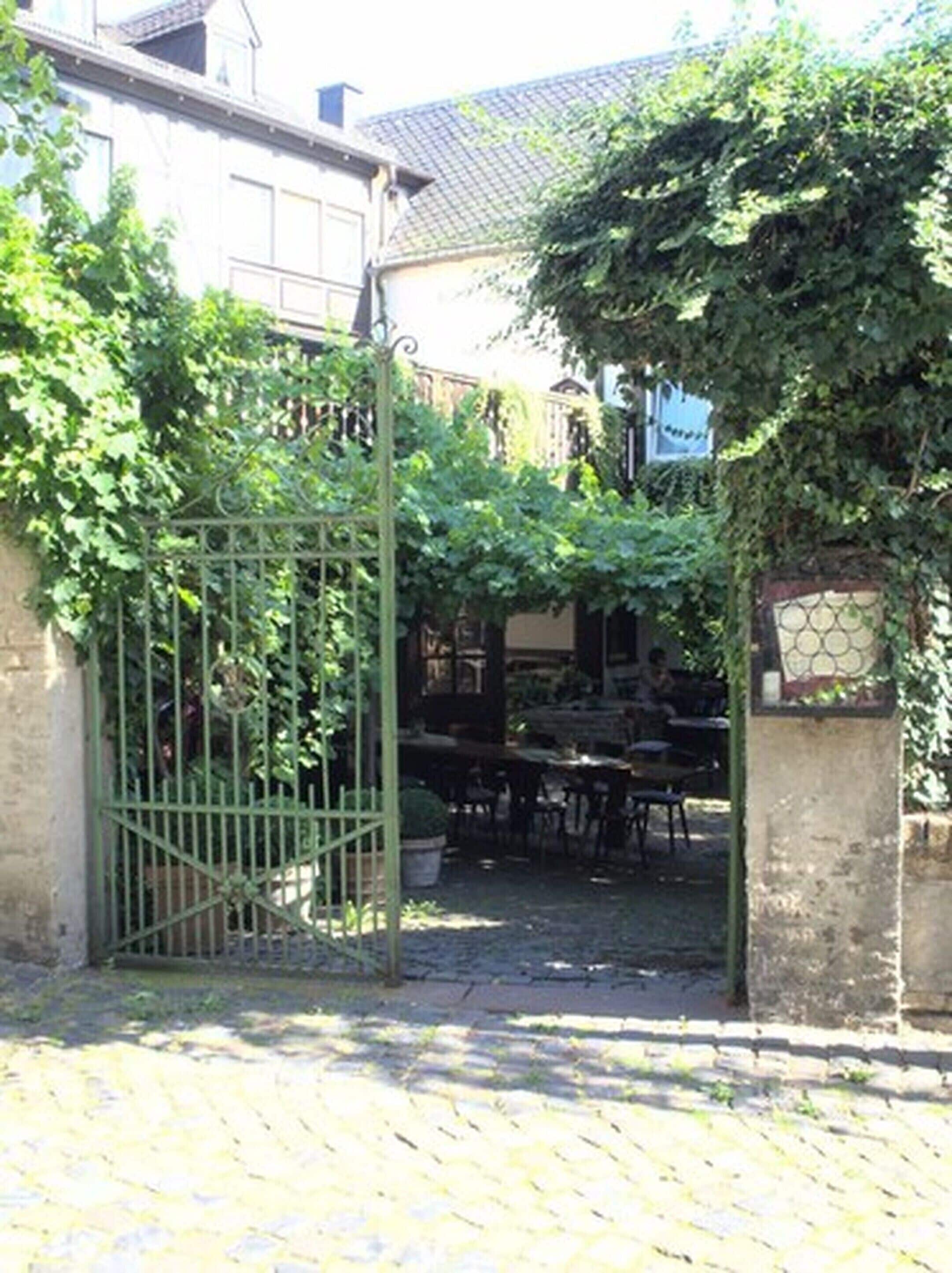 A courtyard with a gate, tables, and chairs, surrounded by greenery and a building with a tiled roof.