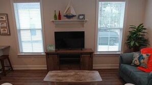 Un salon avec un téléviseur posé sur un meuble en bois, une petite table, un canapé et une fenêtre avec des stores.