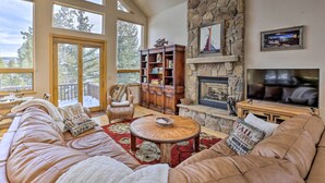 A spacious living room with a stone fireplace, large windows, a cozy sofa, and a bookshelf. - Ski-In/Out Breckenridge Gem w/ Hot Tub! (Breckenridge)
