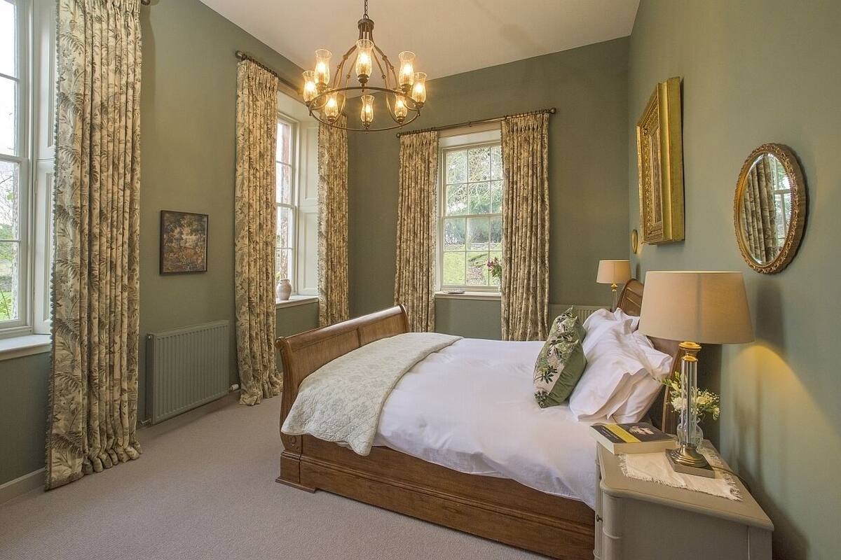 Un dormitorio con una cama de madera, una mesita de noche con lámpara, un espejo y cortinas.