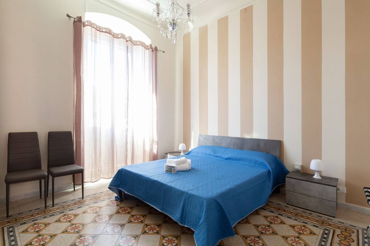 Un dormitori amb llit, dues cadires, una taula de nit, una làmpada de cristall i una finestra amb cortines.