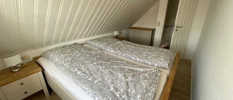 Ein Schlafzimmer mit schrägverdeckter Decke, einem Doppelbett mit gemusterten Betttextilien, einem Nachttisch und einer kleinen Kommode.