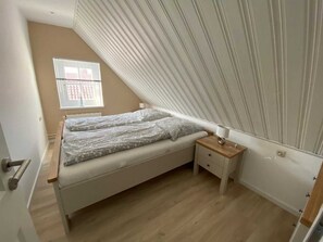 A bedroom with a sloped ceiling, a bed with a mattress and pillows, a bedside table with a lamp, and a window with blinds.