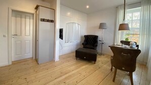 Une pièce spacieuse avec un plancher en bois, un fauteuil noir, une table d’appoint avec une lampe et une fenêtre avec des rideaux.