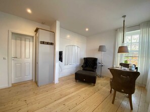 A spacious room with wooden flooring, a black armchair, a side table with a lamp, and a window with curtains.