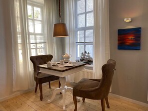A dining area with a table set for two, a ship model on the windowsill, and a painting on the wall.