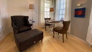 A living room with a dark brown sofa, a matching ottoman, a dining table with chairs, a floor lamp, and a painting on the wall. - Berth 6 - Apartments Meerhuus, 45229 (Jemgum)