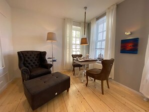 Een woonkamer met een donkerbruine bank, een passende voetenbankje, een eettafel met stoelen, een vloerlamp en een schilderij aan de muur.