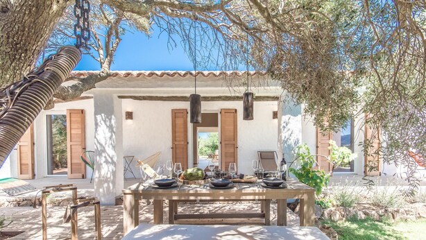 An outdoor dining area with a wooden table set for a meal, surrounded by a white building with wooden shutters and hanging lights.