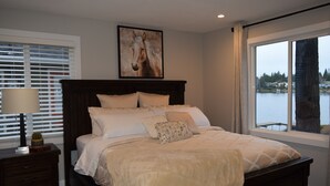 Un dormitorio con una cama grande, una mesita de noche, una lámpara, una ventana con vista a un lago y un cuadro en la pared.