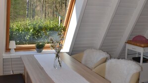 Une pièce mansardée chaleureuse, avec un plafond en pente, une table en bois, une chaise et une vue sur une forêt par une grande fenêtre.