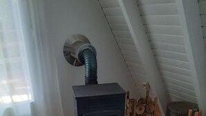 Un poêle à bois dans une pièce dont le sol est recouvert de planches de bois, avec une fenêtre ornée de rideaux et un plafond avec des poutres blanches.