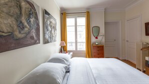 A bedroom with a large bed, a nightstand, a dresser, a window with curtains, a radiator, and a painting on the wall. - Colour in Ternes, by Veeve (Paris)