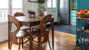 A dining area with a wooden table and chairs, a hanging light fixture, and a kitchen area with a refrigerator and a teal cabinet. - Colour in Ternes, by Veeve (Paris)