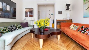A living room with a wooden herringbone floor, a round coffee table with a vase of yellow flowers, an orange sofa, and a green sofa with patterned cushions. - Colour in Ternes, by Veeve (Paris)