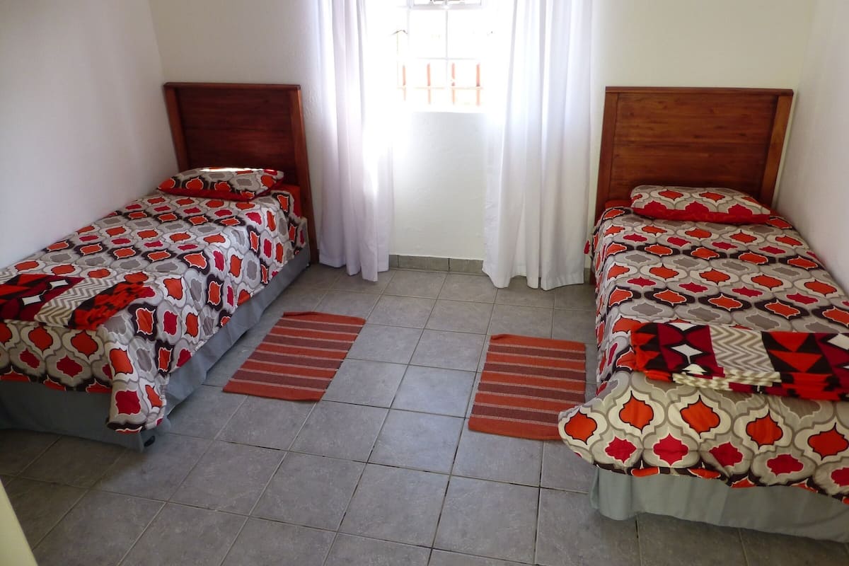 A room with two single beds, each with a patterned bedspread, a wooden headboard, and a window with white curtains.