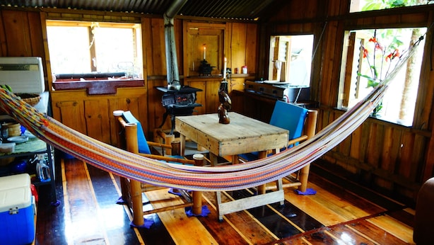 A rustic wooden cabin with a hammock over a wooden table and chairs, a kitchenette, and a window with a view of greenery.