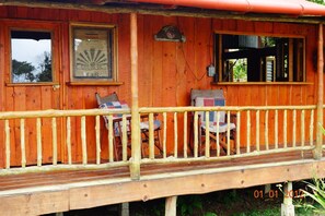 A wooden cabin with a porch, two chairs, a table, and a window with a sign.