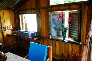 A rustic wooden cabin interior with a dining table, a blue chair, a stove, and a window with a view of greenery.
