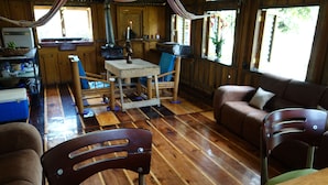 A rustic cabin interior with a wooden floor, a dining table with chairs, a sofa, and a kitchen area with a stove and sink.