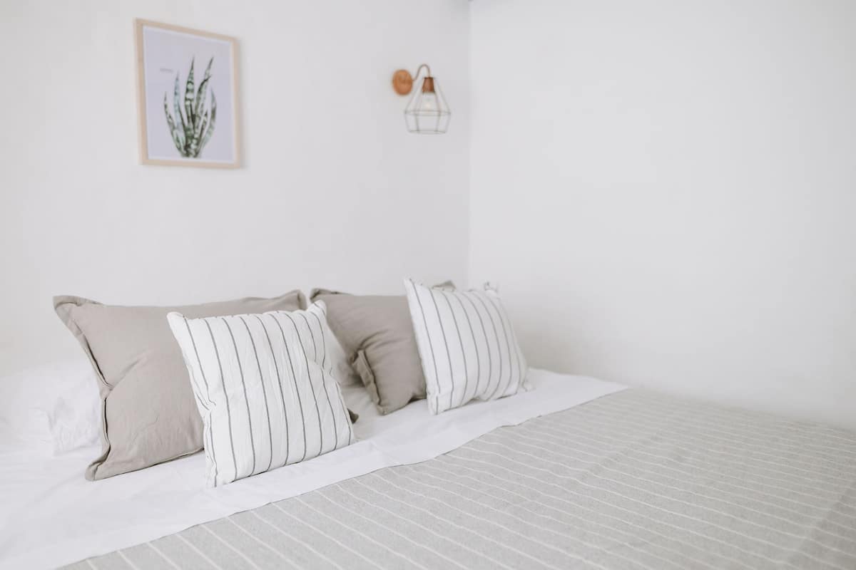 Ein ordentlich bezogenes Bett mit gestreifter Bettwäsche und einer schlichten weißen Wand mit einer gerahmten Pflanze sowie einer Wandleuchte.