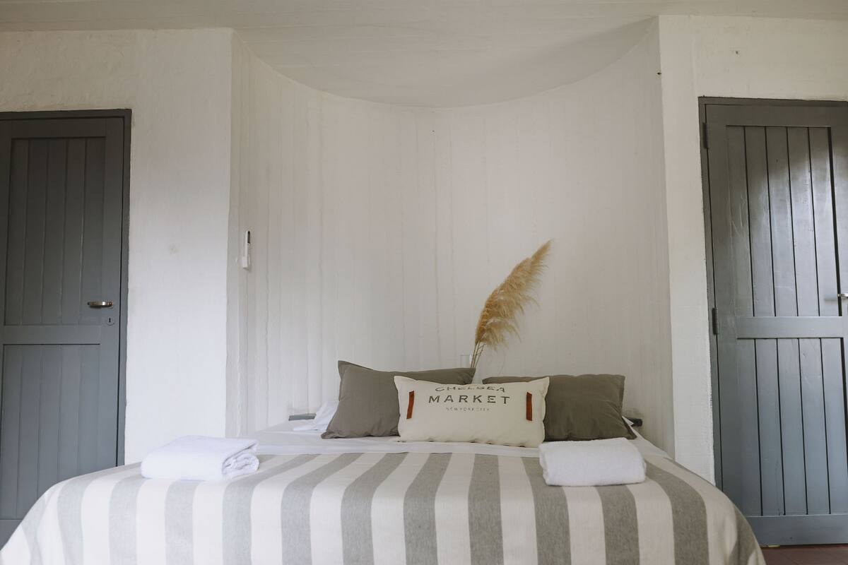 Ein Bett mit gestreifter Bettwäsche, ein Kissen mit der Aufschrift 'Chelsea Market' und eine hohe, getrocknete Pflanze.