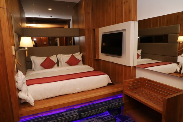 A hotel room with a bed, wooden headboard, a TV mounted above the bed, a mirror on the wall, and a bench at the foot of the bed.
