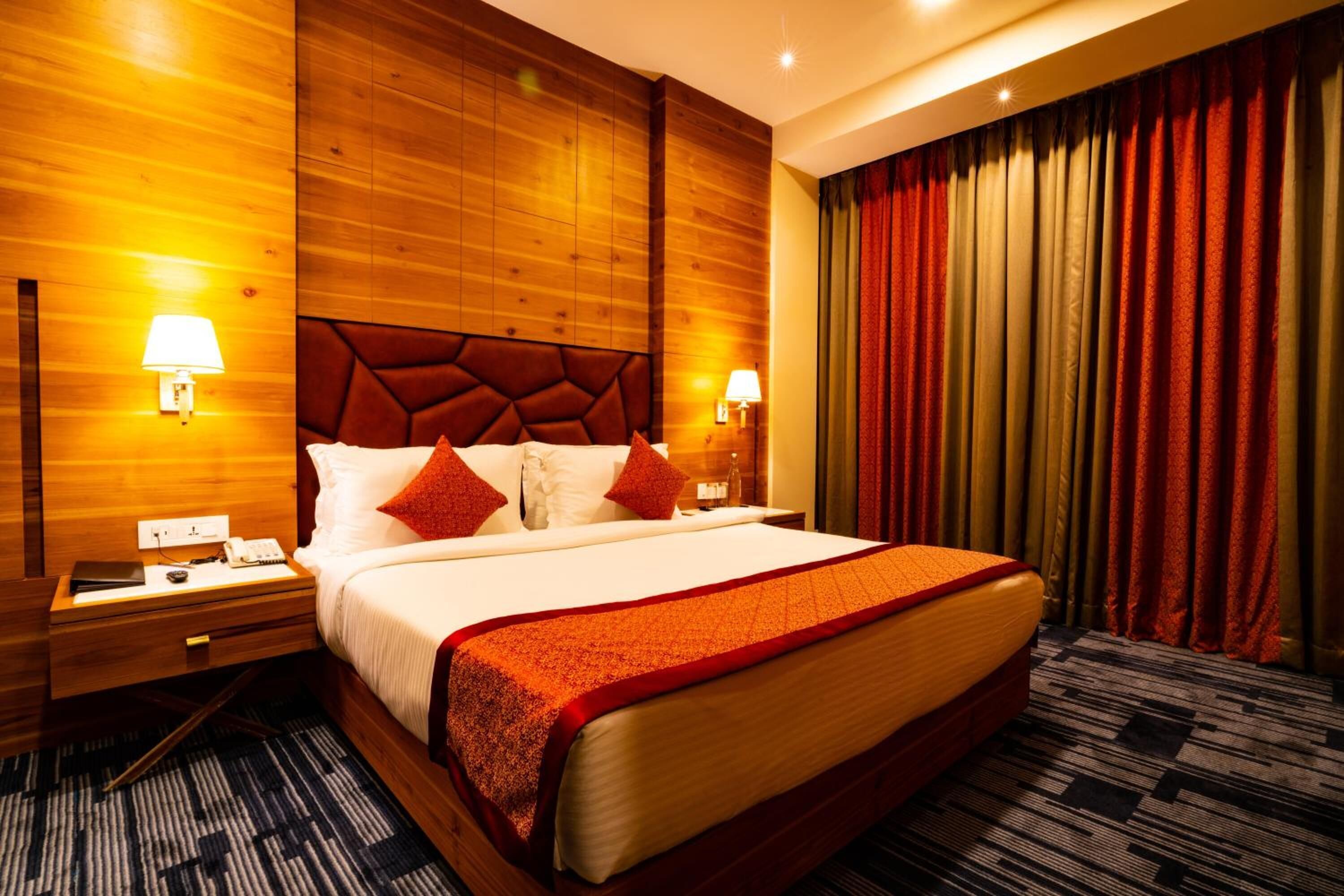 A hotel room with a large bed, wooden panel walls, two bedside lamps, a nightstand, and a striped carpet.