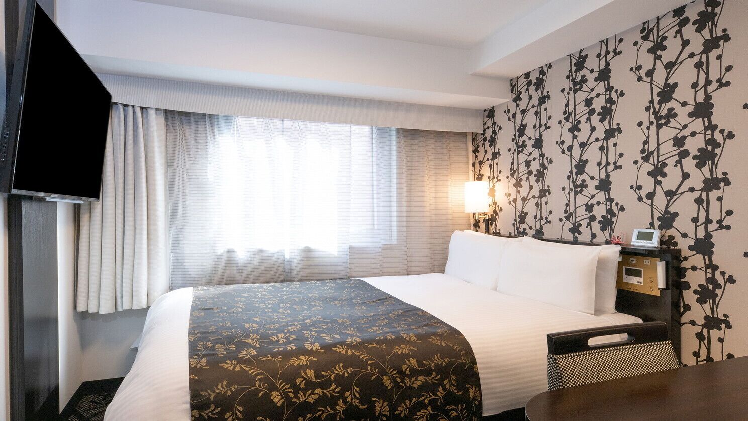 Habitación de hotel con una cama, una mesita de noche con lámpara, un televisor y una pared con estampado floral.