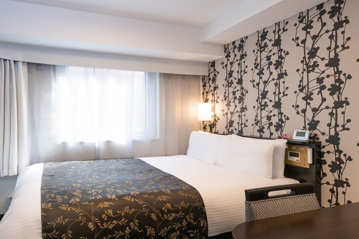Habitació d'hotel amb llit, una taula de nit amb una làmpada, televisió i un mur amb motius florals.