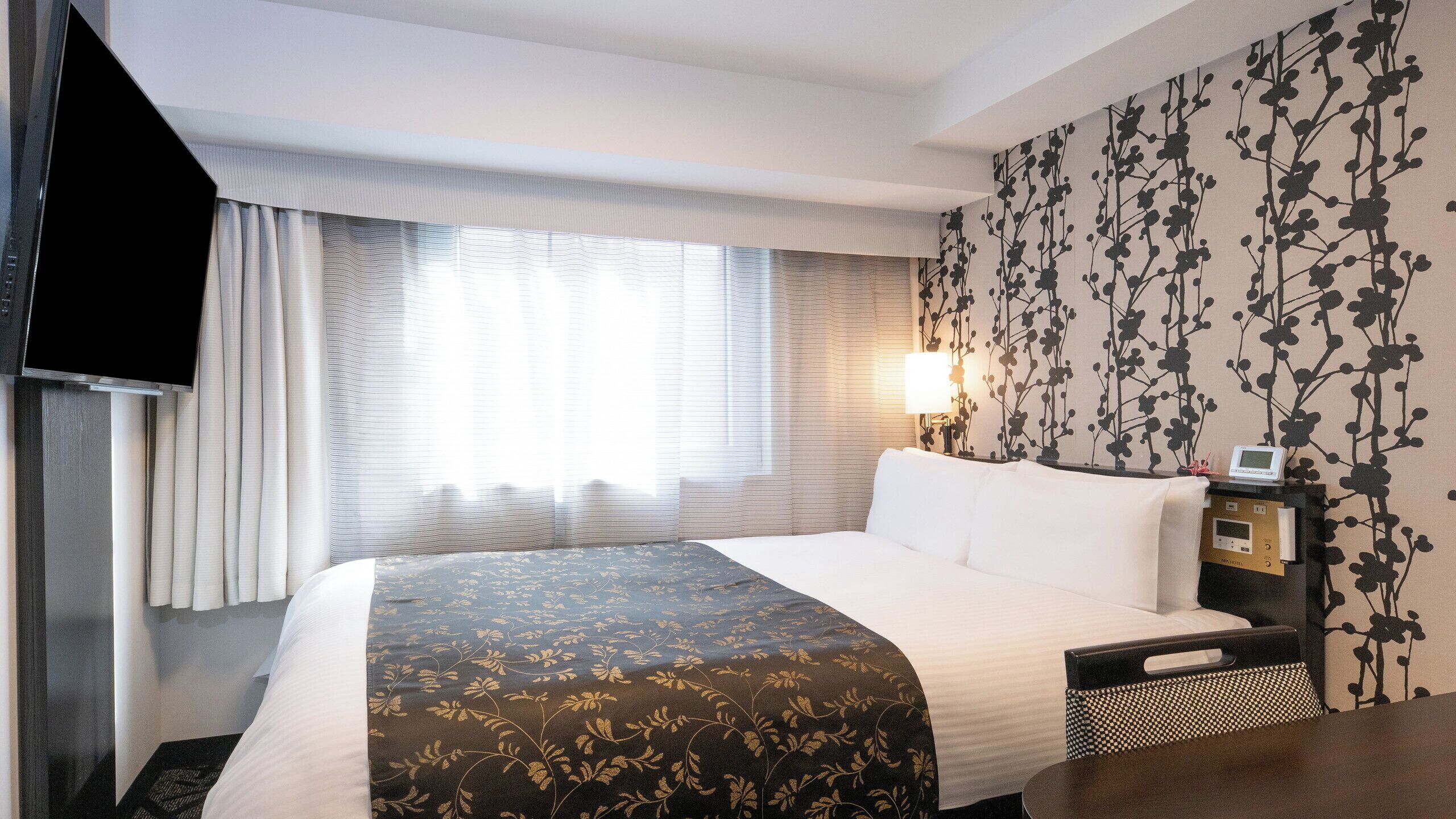 Ein Hotelzimmer mit Bett, Nachttisch mit Uhr, Lampe und Wand mit Blumenmuster.