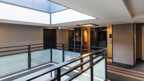 A modern hotel interior with a glass balcony, elevator, and a sign indicating room numbers.