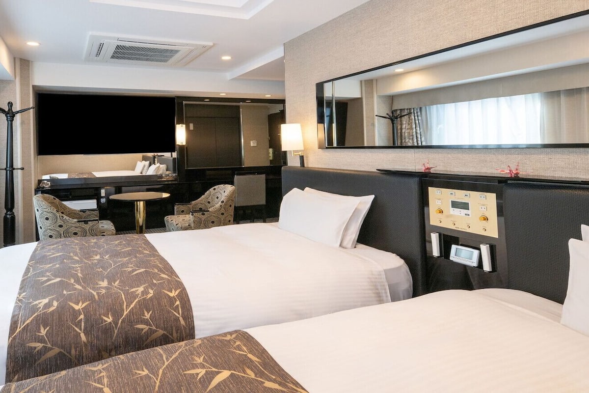 Habitació d'hotel amb dos llits, un escriptori, una cadira, un mirall i un panell de control.