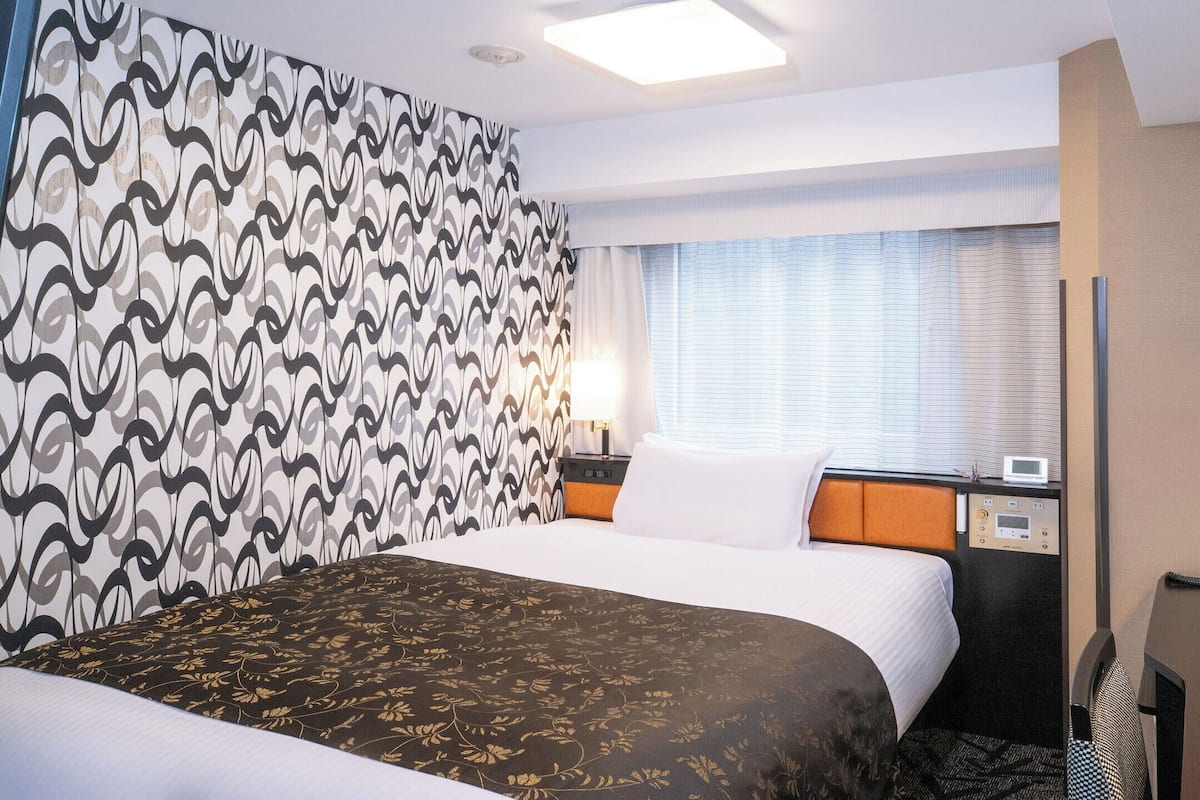 Habitació d'hotel amb llit, taulell de nit, làmpada i panell de control.