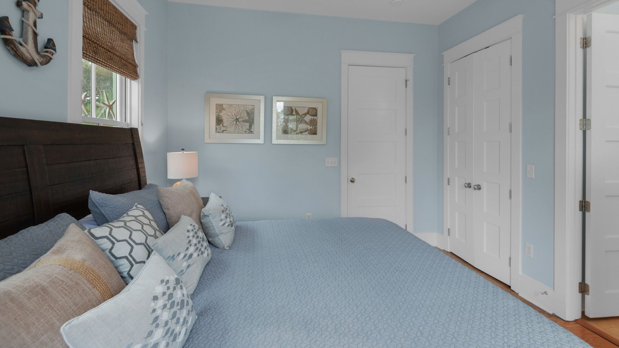 Een slaapkamer met een bed, kussens, een nachtkastje, een lamp en twee foto’s in lijsten aan de muur.
