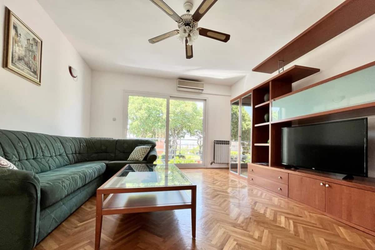 Un salón con un sofá verde, una mesa de centro de vidrio, un mueble de madera con un televisor y un ventilador de techo.