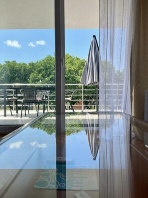 Une table en verre qui reflète un balcon avec des meubles d’extérieur, et d’où l’on peut admirer les arbres et le ciel.