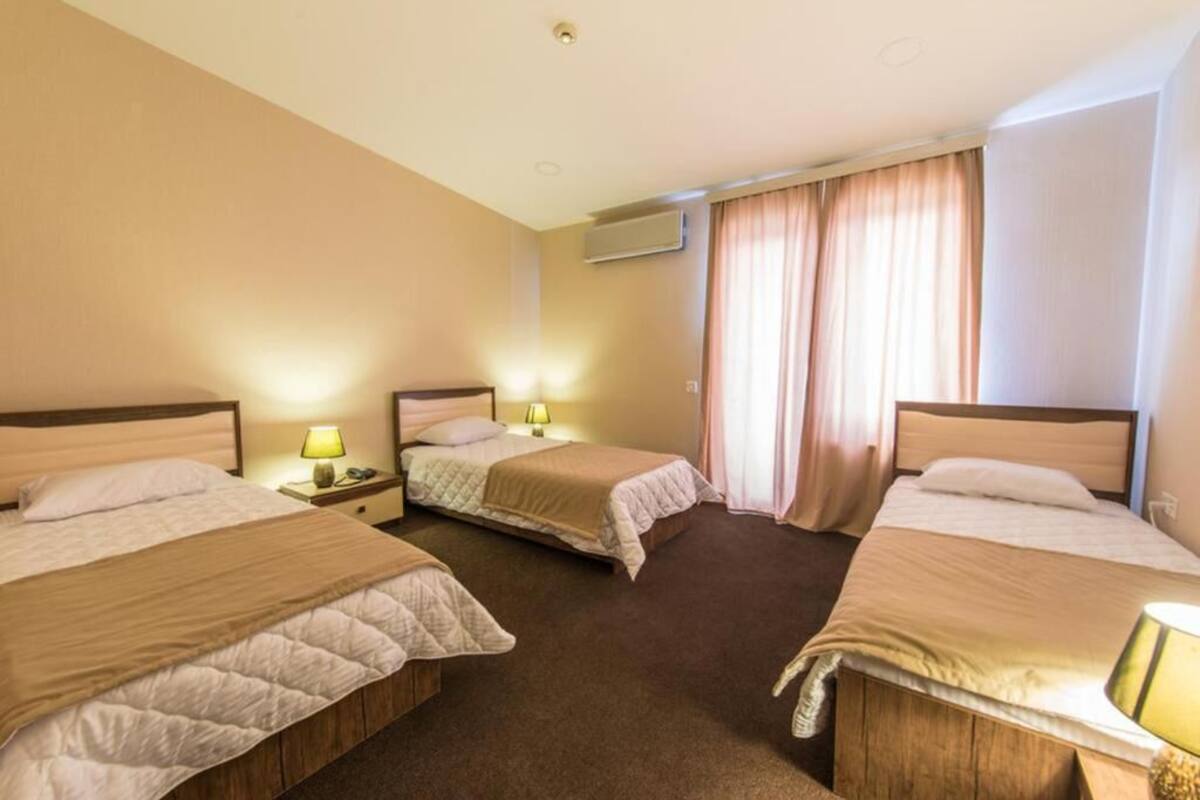 A hotel room with three beds, each with a bedside table and lamp, a window with curtains, and an air conditioning unit on the wall.