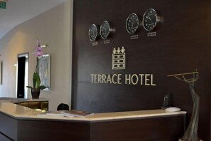 A hotel reception desk with clocks displaying Moscow and New York times, a reception counter, and a plant. - Check-in TERRACE Hotel (Baku)