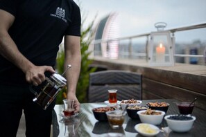 Une personne verse une boisson dans un verre sur une terrasse où une table est dressée avec divers plats et une bougie.