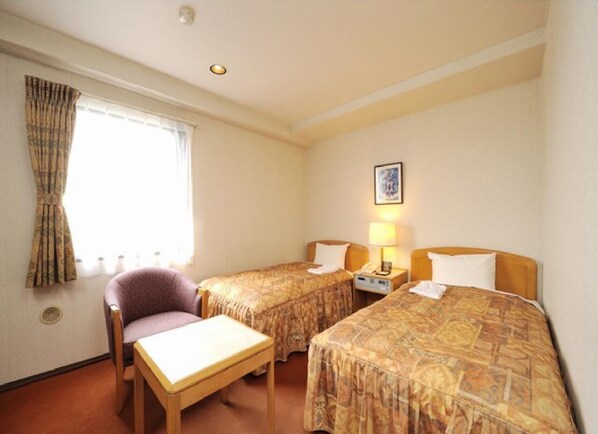 Ein Hotelzimmer mit zwei Einzelbetten, einem kleinen Tisch, einem Stuhl und einem Fenster mit Vorhängen.