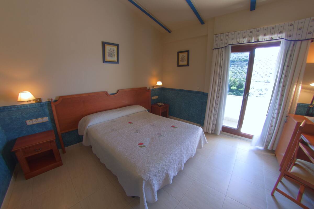 A bedroom with a bed, bedside tables, a window with curtains, and a view of greenery outside.