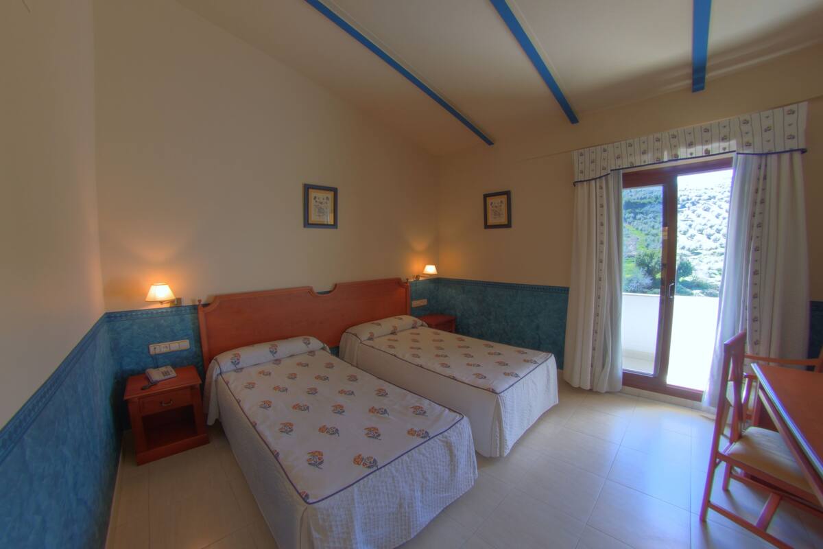 A bedroom with two beds, a wooden nightstand with a lamp, and a view of a garden through a sliding door.