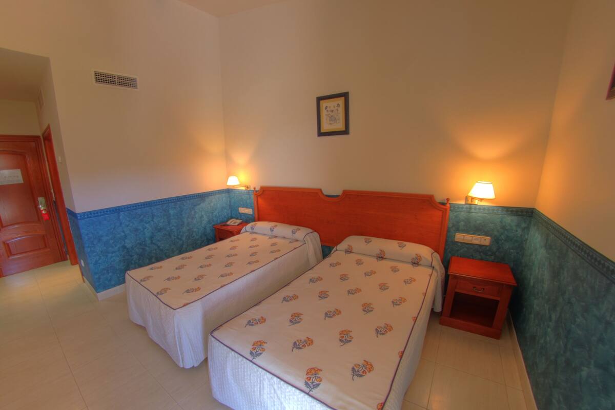 A hotel room with two single beds, a wooden headboard, a nightstand with a lamp, and a framed picture on the wall.