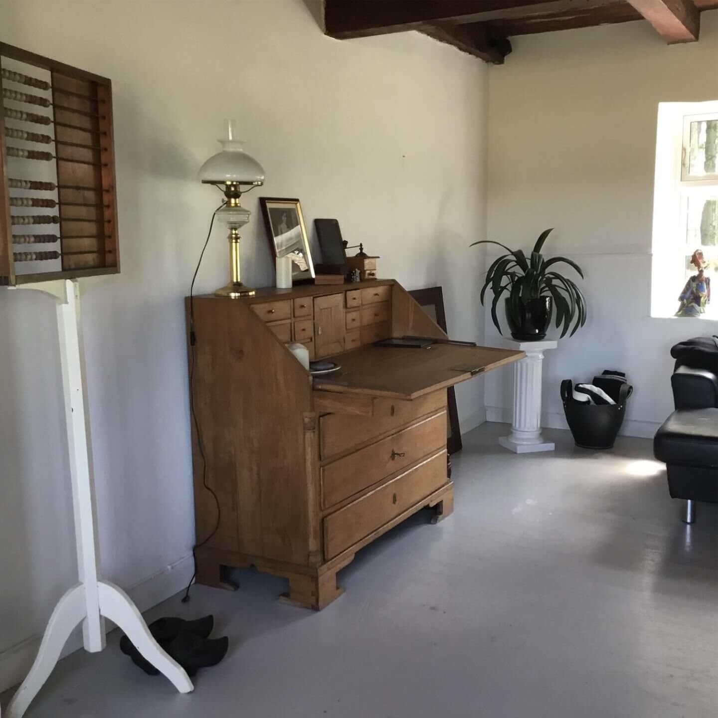 A wooden desk with drawers, a lamp, and a framed photo. There is a potted plant and a black chair in the room.