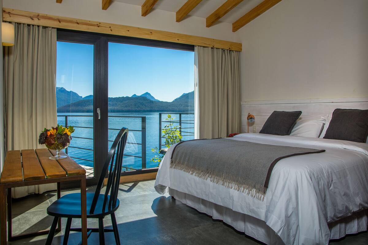 A bedroom with a large bed, a wooden table, a chair, and a view of a lake and mountains through a sliding glass door.