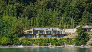 A modern multi-story house with large windows, situated by a lake, surrounded by dense forest. - Correntoso TownHouses (Villa La Angostura)